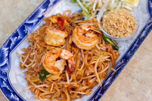 Pad thai noodles with king prawns on a white and blue plate, with peanut butter in a small jar