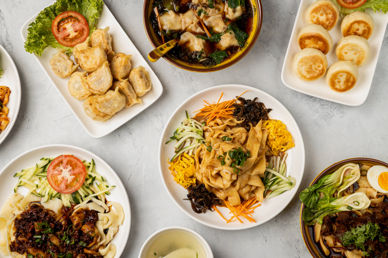 A range of Chinese dishes including five colour noodles, buns and baos, and pan fried dumplings, on a white table.